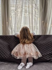 Rear view of girl sitting on sofa at home, looking out the window