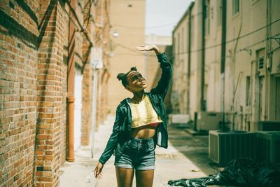 Young woman looking up with arms raised amidst buildings on sunny day