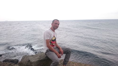 Young man sitting on rock by sea against sky