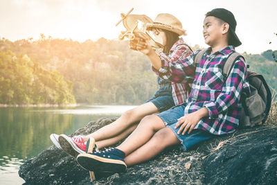 Close-up of friends with cardboard airplane sitting on rock against lake