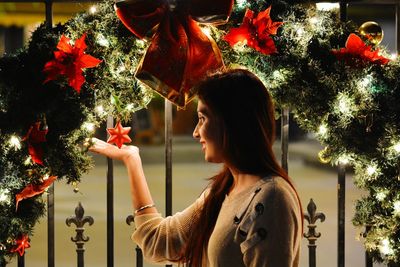 Side view of smiling young woman standing by christmas tree at night