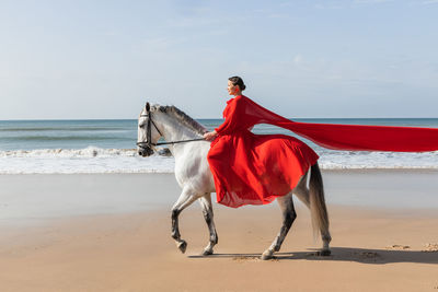 Side view of stylish female in red dress riding purebred horse on coast against stormy ocean