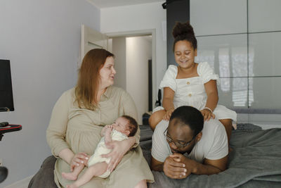 Happy mother looking at daughter sitting on top of her father in bedroom