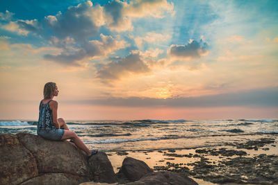 Woman sitting on rock at beach against sky during sunset