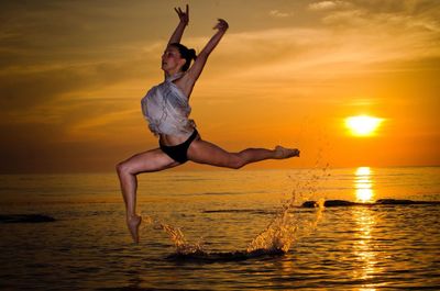 Beautiful woman performing gymnastics in sea against orange sky during sunset