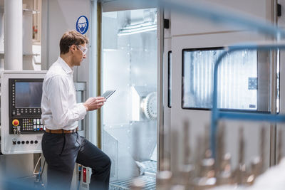 Businessman at machine in factory wearing safety goggles holding laptop