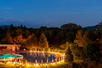 Illuminated swimming pool by lake against sky at night