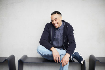 Happy male student looking down while sitting on bench against wall in university