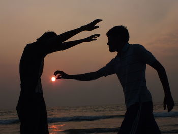 Optical illusion of man holding sun by friend on shore during sunset