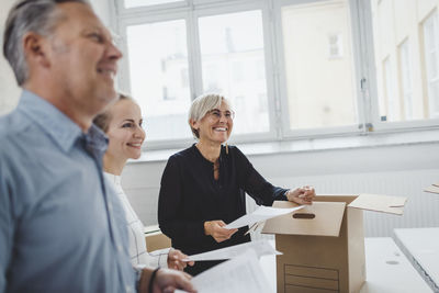 Smiling mature businessman with colleague unpacking cardboard box in new office