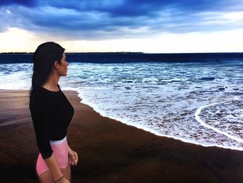 Young woman standing at beach against cloudy sky