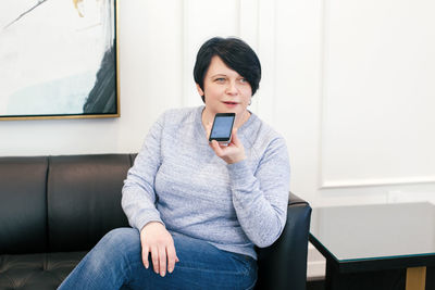 Middle age brunette woman sitting on couch sofa at home and talking on audio chat phone with friends