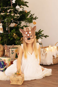 Portrait of cute girl wearing mask sitting against christmas tree at home