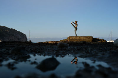 Full body side view of unrecognizable female in sportswear performing standing hand to toe yoga pose during practice on rocky sea coast