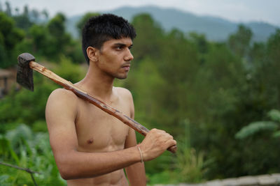 Shirtless young man holding axe standing outdoors