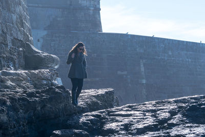 Young woman standing on rock formation by historic building at cascais