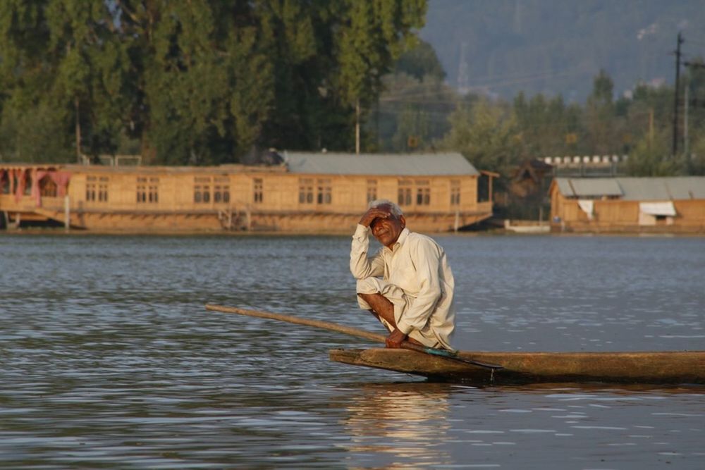 water, real people, nautical vessel, river, one person, waterfront, built structure, outdoors, architecture, senior men, building exterior, transportation, day, mode of transport, men, nature, oar, sailing, leisure activity, full length, tree, fisherman, lifestyles, rowing, beauty in nature, people