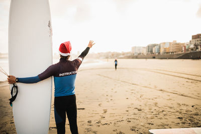 Rear view of man wearing santa hat holding surfboard at beach