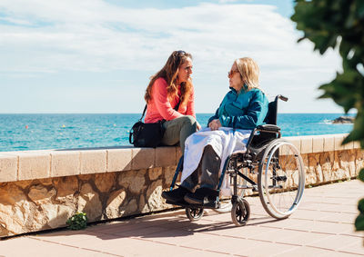 Adult woman and aged mother in wheelchair chilling on embankment and talking to each other on sunny day near sea