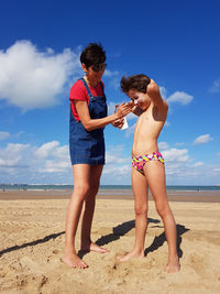 Full length of mother applying moisturizer to daughter at beach during sunny day
