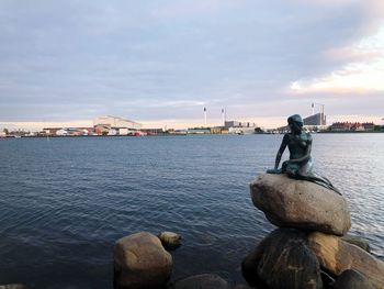 Man sitting on rock by river against sky during sunset