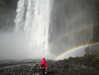 Rear view of man standing by waterfall against rainbow in sky