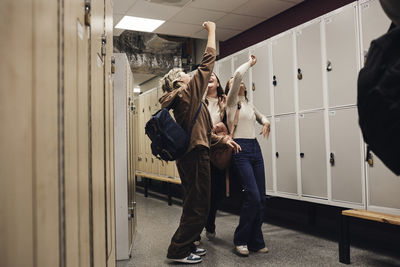 Full length of cheerful female students with hands raised enjoying in school corridor