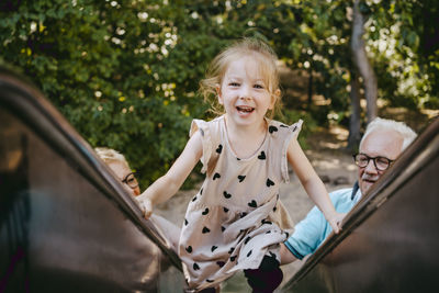 Portrait of smiling girl climbing on slide while playing with grandparents at park