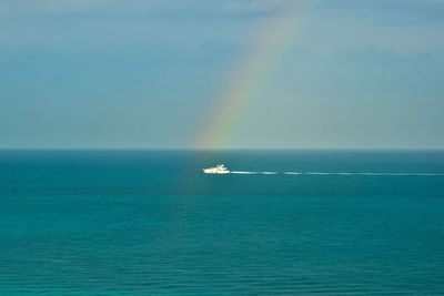 Lone white ship on azure waters at the end of a brilliant rainbow.