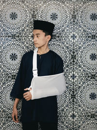 Portrait of a young malay man with injured arm standing against a decorative wall