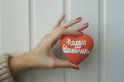 Cropped image of woman holding orange heart shape gingerbread with text against white door