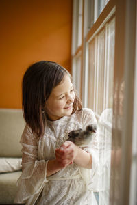 A smiling little girl holds baby chicken up to the window to look out