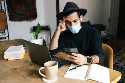 Pensive ethnic male freelancer wearing medical mask sitting at table in home office and browsing smartphone while working during coronavirus pandemic