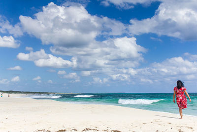 Rear view of mid adult woman walking at beach against cloudy sky