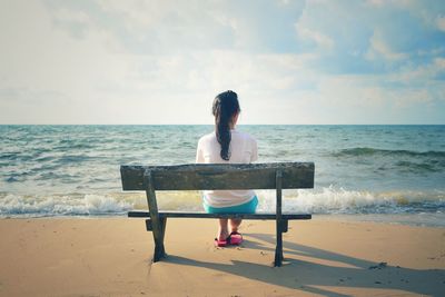 Rear view of woman sitting on bench at beach against sky