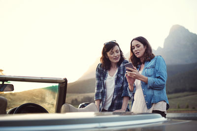 Woman showing mobile phone to friend by convertible car during sunset