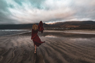 Rear view of woman walking at beach against cloudy sky