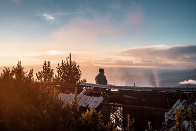 Rear view of man sitting on roof against sky during sunset