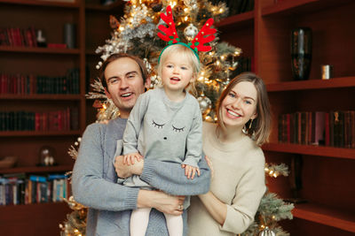 Smiling caucasian mother and father with baby girl playing together by decorated christmas tree