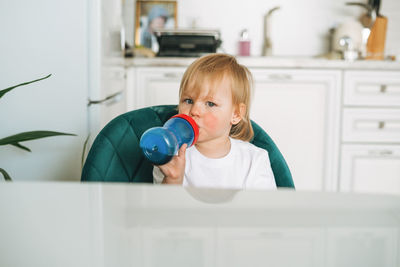 Cute baby girl in home clothes drinking water from bottle in kitchen at home