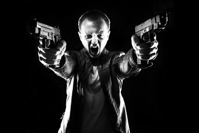 Angry man aiming with guns while standing against black background