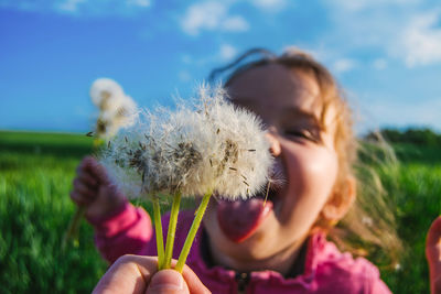 Cropped hand holding dandelions by playful girl against sky