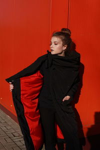 Fashionable teenage girl wearing black warm clothing standing against red wall during sunny day