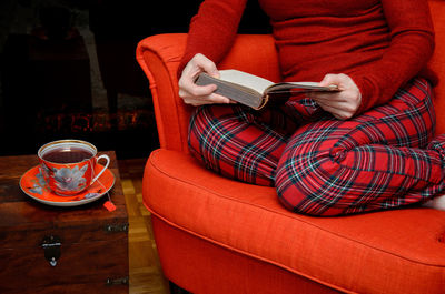 Lady sitting in an armchair in cozy homeware, reading a book and drinking tea by the fireplace