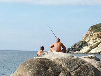 Rear view of shirtless man on rock by sea against sky
