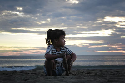 Boy sitting on shore at beach against sky during sunset