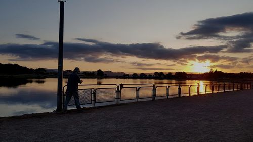 Silhouette man standing by railing on lake against sky during sunset