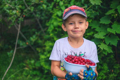 Portrait of cute boy holding red fruits in bowl against plants