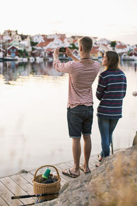 Rear view of couple photographing view through mobile phone by lake