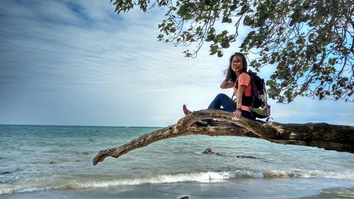Portrait of smiling woman sitting on branch at beach against sky